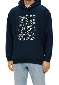Hombre vistiendo una sudadera con capucha azul marino y un diseño abstracto en blanco en el frente, junto con unos jeans azul claro, estilo casual.