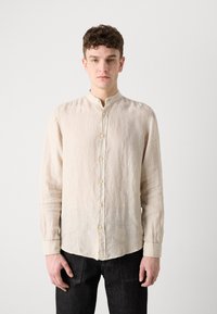 Camicia button-up beige in lino con colletto alla cinese, caratterizzata da una finitura testurizzata e otto bottoni frontali. Indossata con pantaloni in denim scuri.