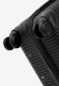 Czarny róg twardej walizki z podwójnymi obrotowymi kółkami i widocznym zamkiem błyskawicznym na jednolitym jasnym tle.