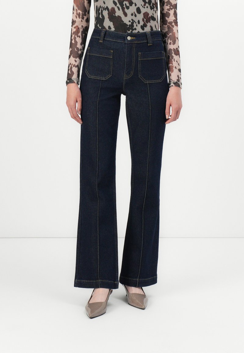 Jean flare bleu foncé avec des coutures en contraste, deux poches arrière et une taille haute. Le tissu est en denim avec une texture lisse.