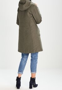 Abrigo largo de color verde oliva con capucha, diseñado con un corte recto y detalles en los bolsillos traseros. Llevado sobre unos vaqueros azules con puños y botas negras de tobillo.