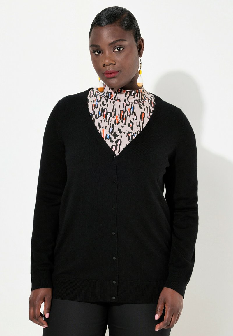 Svart cardigan med V-ringning och knäppning, tillverkad av mjukt tyg. Har långa ärmar och en slät textur, i kombination med en mönstrad topp.