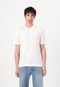 Hvid strikket polo shirt med korte ærmer, der har en ribbet tekstur og knaplukning med tre knapper, parret med lyseblå jeans.