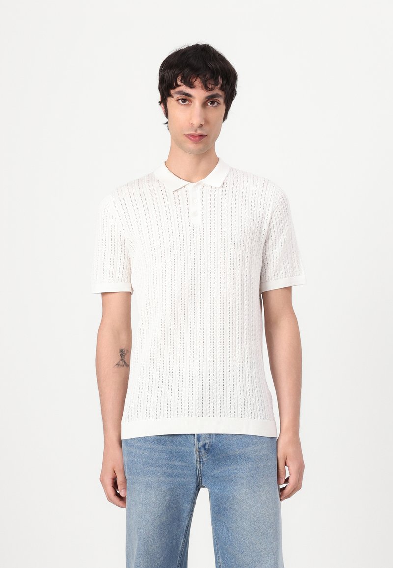 Hvid strikket polo shirt med korte ærmer, der har en ribbet tekstur og knaplukning med tre knapper, parret med lyseblå jeans.