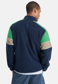 Ζακέτα από φλίς με ναυτικό μπλε βάση, πράσινες και μπεζ λεπτομέρειες, υψηλό γιακά και σχεδιασμό με φερμουάρ στη μέση, προσφέροντας υφή στην εμφάνιση.