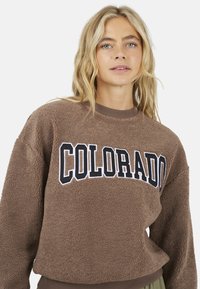 Donna bionda che indossa una felpa marrone in pelliccia sintetica con la scritta "COLORADO" stampata in grandi lettere nere e bianche sul petto.