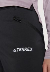 Os pantalões Terrex pretos apresentam um bolso com fecho de correr na lateral, com uma marcação branca na perna inferior e um tecido texturizado para durabilidade.