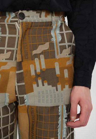 Mønstrede bukser i en blanding af brune, orange og blå farver. Stoffet viser et rutenetdesign med slidte kanter og geometriske former.