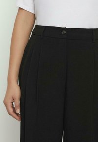 Pantalones de vestir negros con diseño plisado, cierre de botón y bolsillos laterales. La tela tiene una textura suave y un ajuste entallado.