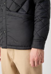 Veste noire matelassée avec boutons et logo "HUGO" sur le bas, portée avec un pantalon beige et une main détendue sur le côté.