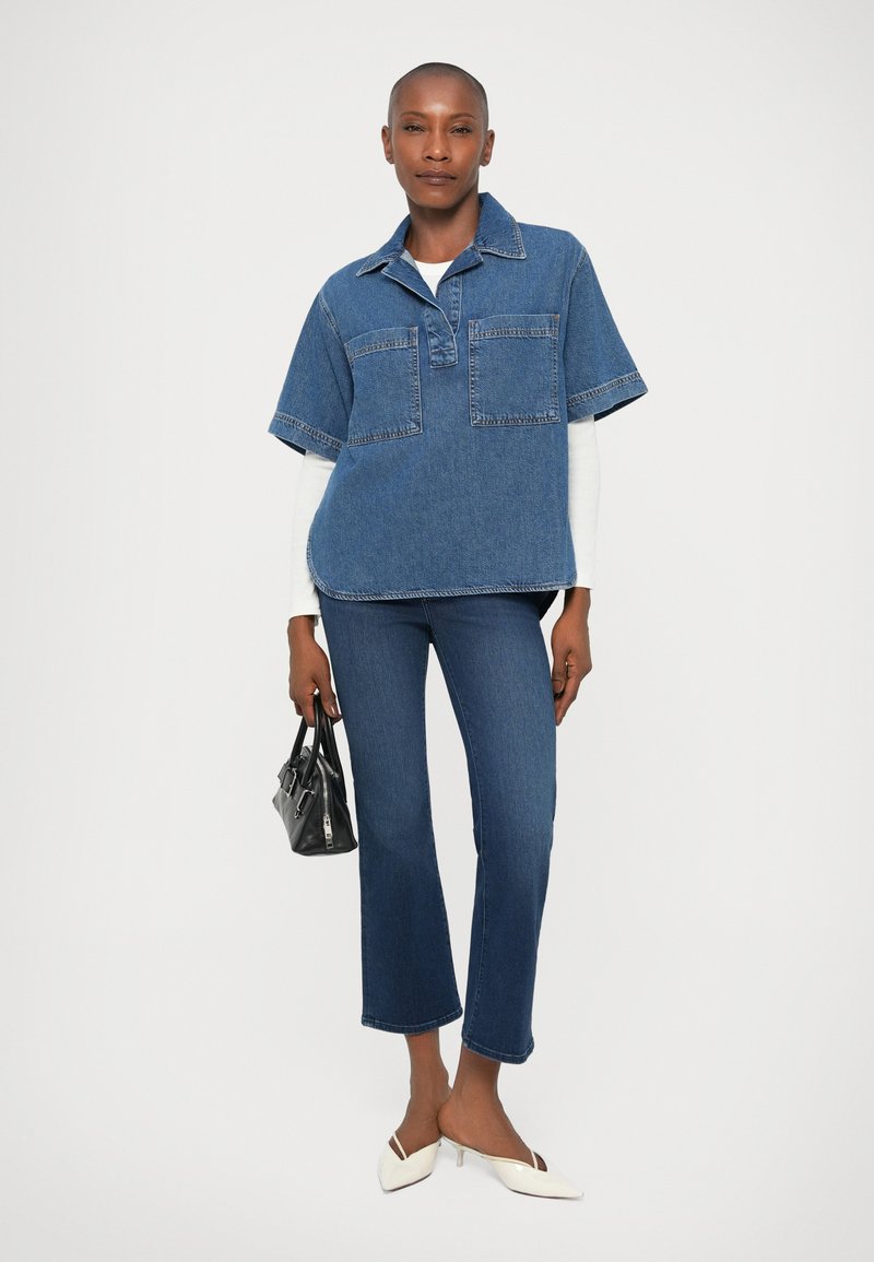 Übergroßes Jeanshemd in Blau mit zwei Brusttaschen; kombiniert mit blauen eng anliegenden Jeans und weißen spitzen Schuhen. Hält eine kleine schwarze Handtasche.
