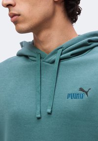 Νεαρό άτομο με τυρκουάζ φούτερ Puma με κορδόνια, δείχνει το πλάγιο προφίλ από τον λαιμό έως το άνω μέρος του στήθους.