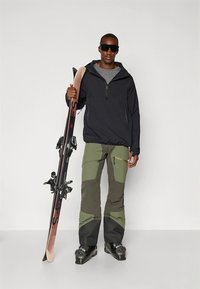 Equipamento de esqui composto por um casaco preto com fecho de correr, camisa cinza, calças de esqui verdes com reforços nos joelhos e botas de esqui pretas, segurando um par de esquis.