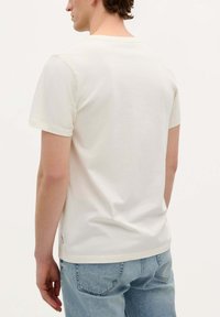 Tricou alb din bumbac, cu guler rotund clasic și mâneci scurte, având o croială dreaptă. Purtați cu blugi albaștri deschiși.