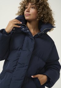 Chaqueta de plumas azul marino con un alto cuello, cremallera frontal, botones a presión y bolsillos laterales. Presenta una textura acolchada con un diseño estructurado.