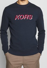 Námořnická modrá mikina vyrobená z měkké látky, s odvážně vyšitým logo "KORS" v červené a bílé barvě na hrudi. Klasický střih s žebrovanými manžetami.