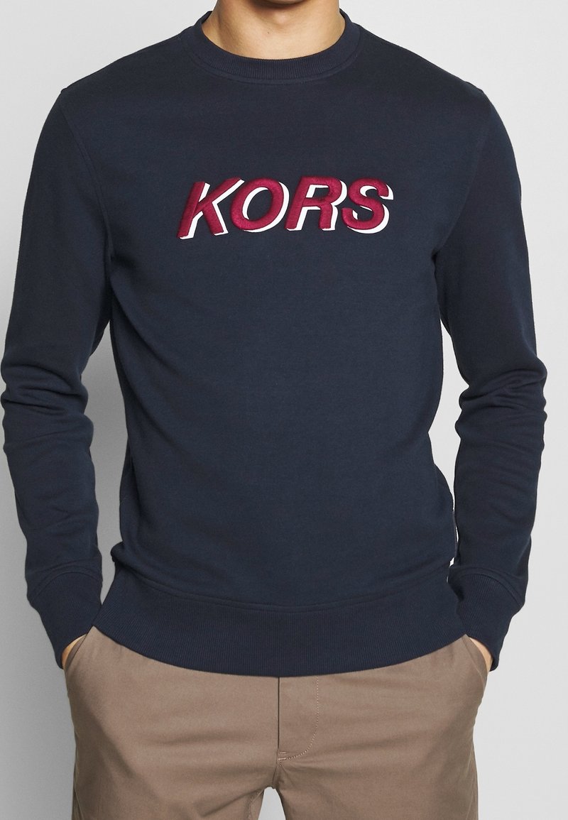 Námořnická modrá mikina vyrobená z měkké látky, s odvážně vyšitým logo "KORS" v červené a bílé barvě na hrudi. Klasický střih s žebrovanými manžetami.