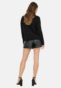 Schwarzer Strickpullover mit rundem Ausschnitt und geripptem Saum, kombiniert mit glänzenden schwarzen Leder-Shorts und klobigen Sandalen mit Absatz.