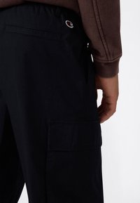 Person in schwarzen Cargo-Hosen mit einer Seitentasche und einem kleinen Champion-Logo in der Nähe des Bundes, sowie einem braunen Langarmshirt.