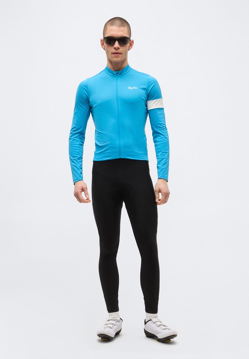 Young man wearing blue long-sleeve cycling jersey, black tights, white cycling shoes, and black sunglasses, standing against plain background.