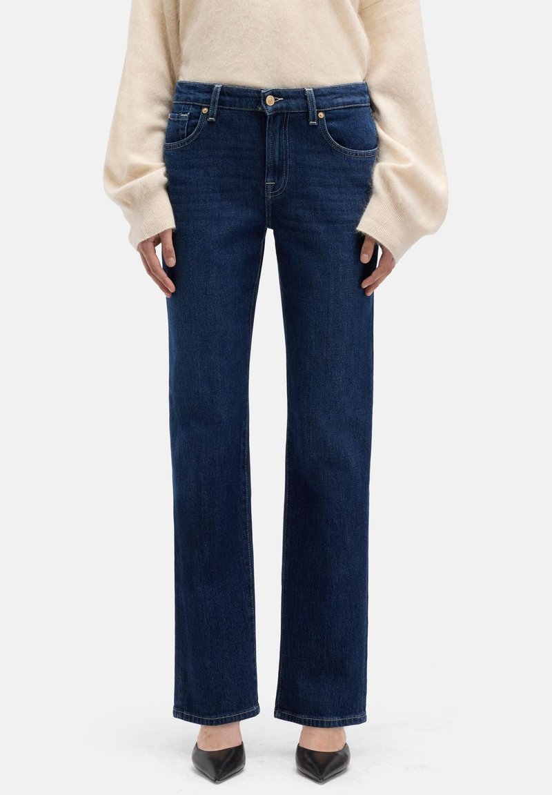 Jeans in denim blu scuro con design a gamba dritta, chiusura con bottone e zip, e tasche frontali. Indossati con un maglione crema e tacchi neri.