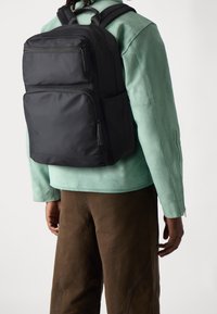 Černý batoh s více zipovými přihrádkami, matným povrchem, nastavitelnými ramenními popruhy a obdélníkovým tvarem, nošený přes zelenou bundu.