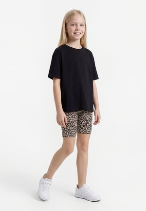 Lachend kind staand, draagt een zwart oversized t-shirt, korte broek met luipaardprint en witte sneakers tegen een witte achtergrond.