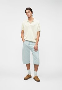 Ανοιχτό κίτρινο ριγέ πόλο πουλόβερ συνδυασμένο με ανοιχτό μπλε τζιν σορτσάκι. Καφέ παπούτσια και λευκές κάλτσες ολοκληρώνουν την εμφάνιση, με μινιμαλιστικό σχέδιο.