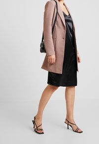 Różowa marynarka, czarna satynowa sukienka na ramiączkach, czarne sandały na obcasie. Marynarka ma strukturę, sukienka gładką fakturę, a sandały mają obcas.