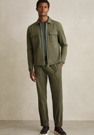 Mann trägt eine olivgrüne Jacke mit Reißverschluss und Klapptaschen, passende Hose, ein dunkelgrünes Hemd und schwarze Turnschuhe und steht vor einer schlichten Wand.