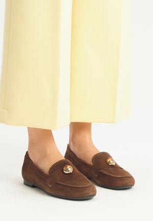 Brown suede loafers with a flat sole, featuring a gold accent on the upper. Worn with yellow wide-legged trousers.