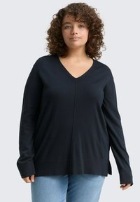 Zwarte lange mouwen V-hals trui met geribbelde textuur en zijsplitten, gemaakt van een zachte stof. Gecombineerd met lichtblauwe jeans.