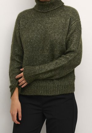 Osoba ubrana w zielony prążkowany golf z długimi rękawami i czarne spodnie, stojąca na tle jednolitego białego tła.