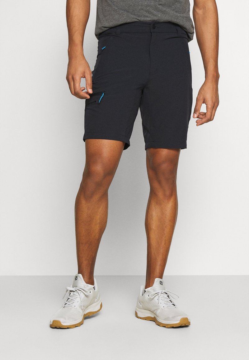 Shorts noirs pour hommes en tissu lisse avec un design ajusté, comportant des poches zippées et des accents bleus sur les côtés. Portés avec des baskets blanches.