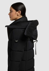 Čierna puffa bunda s prešívaným dizajnom, golierom z umelej kožušiny, zipsovým zapínaním a logom na rukáve. Hladká textúra a dostatočná izolácia.