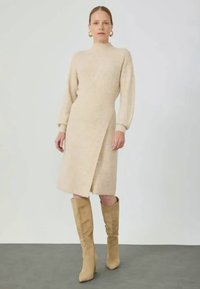 Robe tricotée beige à manches longues et col montant, avec un design enveloppant. Portée avec des bottes taupe hauteur genou sur un sol gris.
