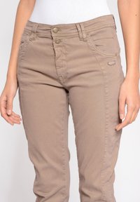 Krémové elastické džíny s přiléhavým střihem, dvěma knoflíkovými zapínáními, předními kapsami a malým logem na přední kapse. Hladká textura.