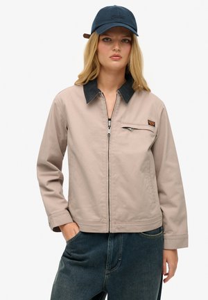 Lichtbeige jas met een donkere corduroy-kraag, rits aan de voorkant en borstzak. Draagt het met donkerblauwe denim voor een casual outfit.