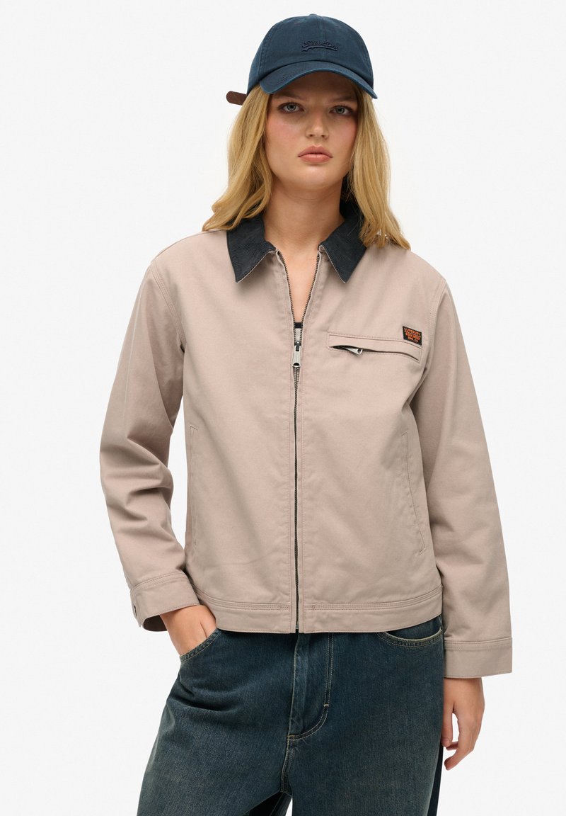 Helle beigefarbene Jacke mit einem dunklen Cordkragen, Frontreißverschluss und Brusttasche. Tragen mit dunkelblauer Jeans vervollständigt ein lässiges Outfit.