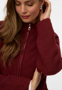 Burgundski zip-up pulover s rebrastim manžetama i teksturom tijela. Srebrni patentni zatvarač i ovratnik, izražen mekim, toplim materijalom.