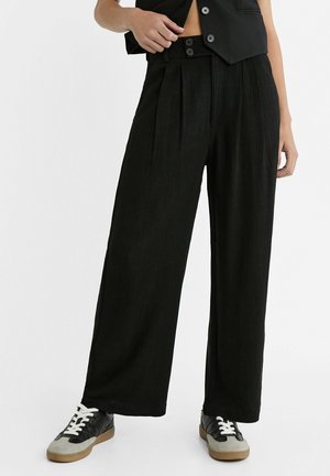 Pantaloni larghi neri con pieghe, dotati di vita alta e chiusura a due bottoni. Indossati con sneakers nere e grigie. Tessuto dalla superficie liscia.