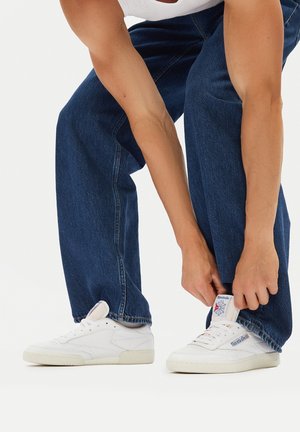 Adidași albi din piele texturată, cu un design cu șireturi și eticheta din limbă cu marcă. Purtați cu blugi din denim decolorat, largi, de culoare bleumarin.