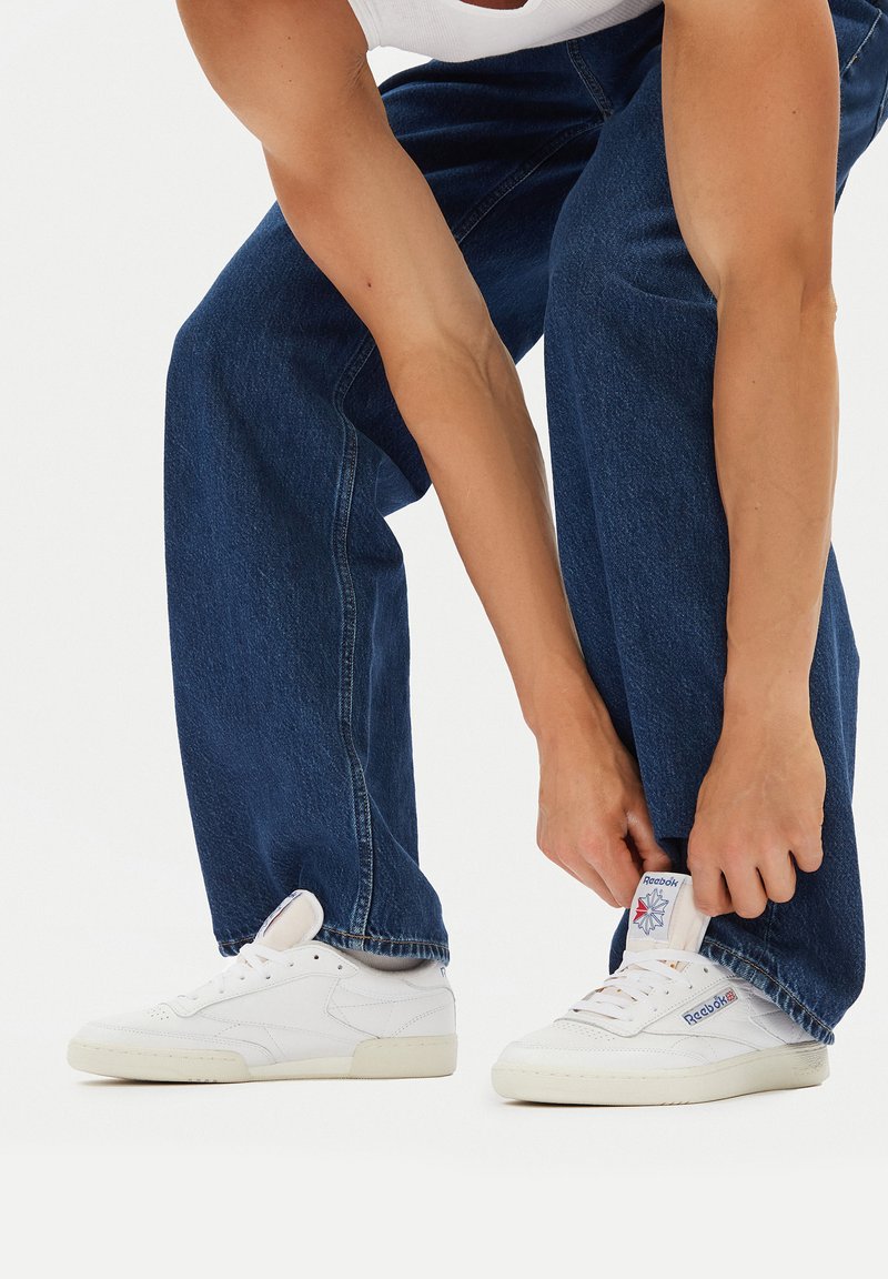 Adidași albi din piele texturată, cu un design cu șireturi și eticheta din limbă cu marcă. Purtați cu blugi din denim decolorat, largi, de culoare bleumarin.