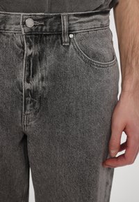 Dark gray denim jeans with a high waist, front pockets, a metal button closure, and a faded texture. Hand visible on the side.