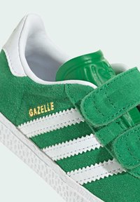 Green suede sneaker with white leather accents, featuring Velcro straps and a textured rubber sole. "GAZELLE" is embossed in gold.