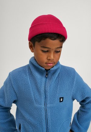 Veste en polaire bleue avec fermeture éclair et col montant. Logo noir sur la poitrine. Bonnet en tricot rose. Garçon regardant vers le bas.
