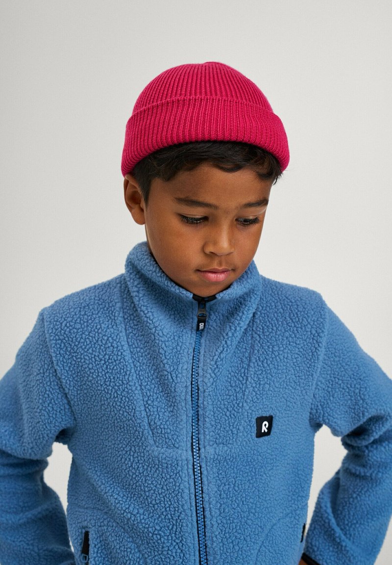 Niebieska polarowa kurtka z zapięciem na zamek i wysokim kołnierzem. Czarny logo na klatce piersiowej. Różowa dziana czapka. Chłopiec patrzy w dół.