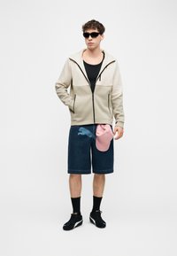 Νεαρός άνδρας φορώντας μπεζ φούτερ με φερμουάρ, μαύρο αμάνικο μπλουζάκι, σκούρες τζιν βερμούδες, μαύρα αθλητικά παπούτσια και γυαλιά ηλίου, κρατώντας ένα ροζ καπέλο στο πλάι του.