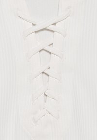 Zblízka bílá žebrovaná látka s křížovými bílými šňůrkami provlečenými očky, které vytvářejí dekorativní zapínání.