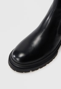 Fekete bőr bokacsizma kerek orral, sima textúrával és vastag gumitalppal, amelyen vízszintes barázdák találhatók a tapadás érdekében.